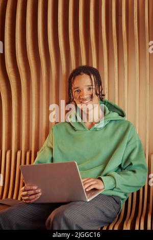 Happy African American teenage guy in green hoodie typing on laptop and looking at camera while sitting by wall in college corridor Stock Photo