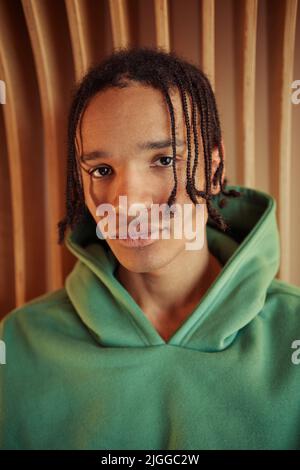 Close-up portrait of serene teenage African American guy in green hoodie looking at camera while standing against beige wall Stock Photo