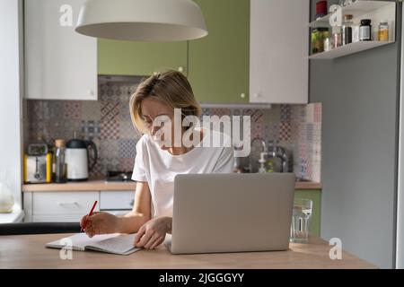 Blonde pensive middle-aged woman working from home using laptop and notebook Stock Photo