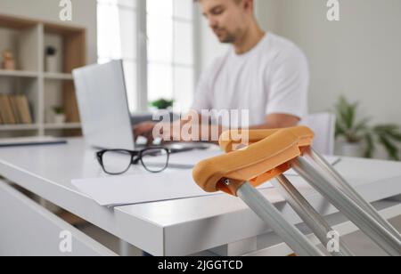 Close up of crutches on background of concentrated man working on laptop in office. Stock Photo