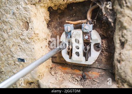A screwdriver unscrews a self-tapping screw in an old disassembled electrical outlet in a clay wall. Dismantling the old outlet Stock Photo