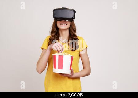 Young smiling female in VR headset watching movie with popcorn, smiling happily, expressing positive emotions, wearing yellow T-shirt. Indoor studio shot isolated on gray background. Stock Photo