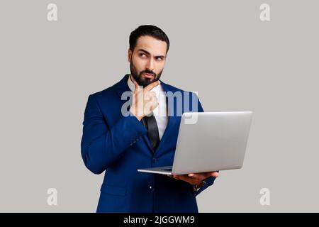 Thoughtful bearded man holding laptop and thinking over startup strategy with serious doubtful expression, wearing official style suit. Indoor studio shot isolated on gray background. Stock Photo