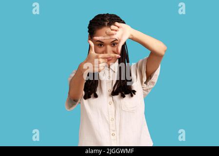 Happy ambitious woman with black dreadlocks makes hand frames, searches perfect angle, smiling, gazes at camera through hands, wearing white shirt. Indoor studio shot isolated on blue background. Stock Photo