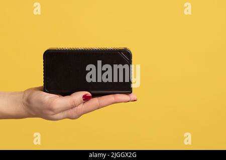 Profile side view closeup of woman hand holding and showing black wireless music speaker, bluetooth musical column. Indoor studio shot isolated on yellow background. Stock Photo