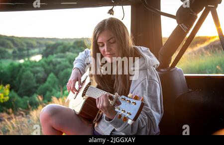 A young woman plays the acoustic guitar in the trunk of a car in nature. Stock Photo