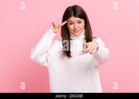 Portrait of beautiful woman holding finger near head and pointing finger at camera, stupid gesture, wearing white casual style sweater. Indoor studio shot isolated on pink background. Stock Photo