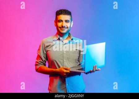 Portrait of happy positive man in shirt holding notebook in hands, looking at camera with positive facial expression. Indoor studio shot isolated on colorful neon light background. Stock Photo