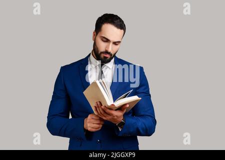Concentrated bearded man reading book, enjoying reading, standing absorbed with exciting plot, checking schedule in notepad, wearing official style suit. Indoor studio shot isolated on gray background Stock Photo