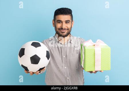 Portrait of satisfied pleased businessman standing looking smiling at camera, holding out soccer ball and present box, wearing striped shirt. Indoor studio shot isolated on blue background. Stock Photo