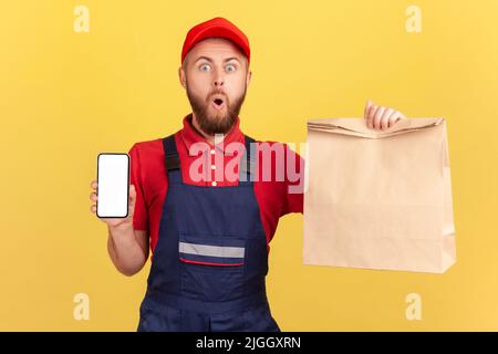 Amazed surprised deliveryman holding paper parcel and showing mobile phone with empty display for advertisement, looking at camera with open mouth. Indoor studio shot isolated on yellow background. Stock Photo