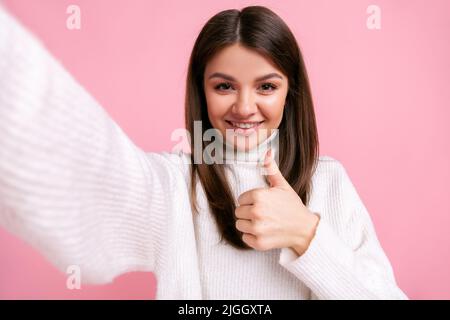 Positive young adult optimistic girl making point of view photo, showing thumb up, approval gesture, wearing white casual style sweater. Indoor studio shot isolated on pink background. Stock Photo