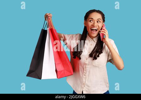 Portrait of excited amazed woman with black dreadlocks holding shopping bags and talking phone, boasting purchase to her friend, wearing white shirt. Indoor studio shot isolated on blue background. Stock Photo