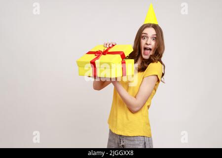 Portrait of excited amazed brown haired young woman unpacking gift box with surprised expression, enjoying unwrapping birthday present. Indoor studio shot isolated on gray background. Stock Photo