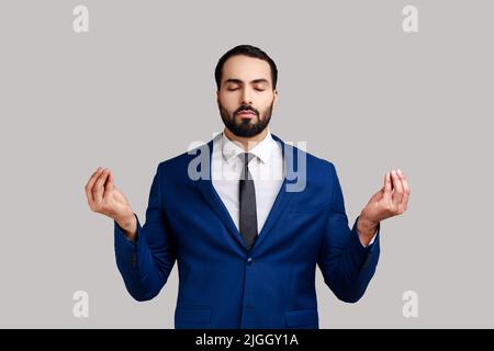 Portrait of relaxed handsome bearded man standing with raised arms and doing yoga meditating exercise, wearing official style suit. Indoor studio shot isolated on gray background. Stock Photo