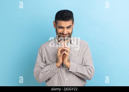 Portrait of funny tricky handsome businessman thinking devious plan with cunning face expression, looking at camera, wearing striped shirt. Indoor studio shot isolated on blue background. Stock Photo