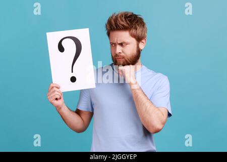 Portrait of puzzled confused bearded man holding white paper with question mark, finding smart solution, asking for advice, holding chin. Indoor studio shot isolated on blue background. Stock Photo