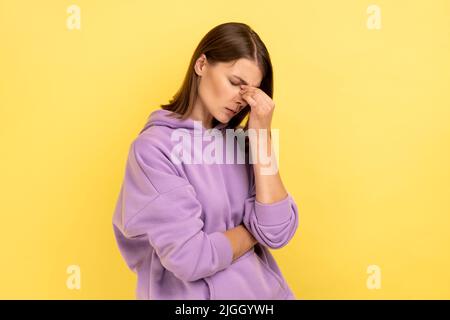 Upset depressed young woman holding her head down, touching face and crying with dramatic grimace, feeling sorrow regret, wearing purple hoodie. Indoor studio shot isolated on yellow background. Stock Photo