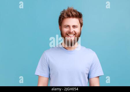 Portrait of bearded man looking at camera with toothy smile and happy facial expression, being in good mood, rejoicing great results. Indoor studio shot isolated on blue background. Stock Photo