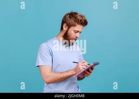 Portrait of bearded man making notes in paper notebook, writing business idea, future plans, checking appointment in schedule diary. Indoor studio shot isolated on blue background. Stock Photo