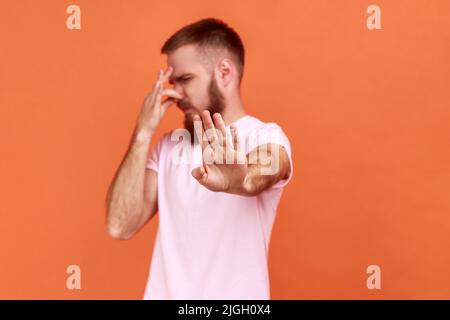 Portrait of bearded man grabbing nose, grimacing in disgust and showing stop gesture, expressing repulsion to stink, fart gases, wearing pink T-shirt. Indoor studio shot isolated on orange background. Stock Photo