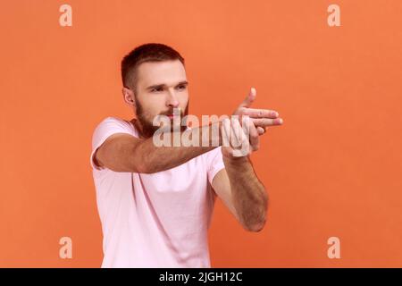 Side view of serious brave bearded man holding fingers pretending holding gun, aiming enemy, self-defense, wearing pink T-shirt. Indoor studio shot isolated on orange background. Stock Photo