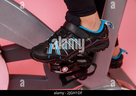 Closeup portrait of the bike simulator and women's legs in black sneakers, cardio workout. Indoor studio shot isolated on pink background. Stock Photo
