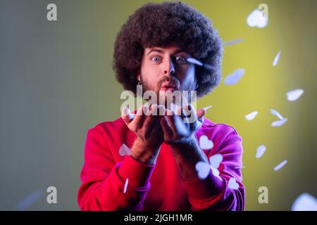 Portrait of hipster man with Afro hairstyle blowing heart shaped confetti, enjoying birthday or valentines day, wearing red sweatshirt. Indoor studio shot isolated on colorful neon light background. Stock Photo