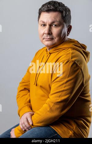 Portrait of confident handsome Caucasian man looking at camera, sitting on chair, looks calm, wearing urban style hoodie. Indoor studio shot isolated on gray background. Stock Photo