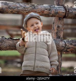 Bully boy kid with a slingshot aims at someone near a fence in the village outdoors. Rustic barefoot child boy with a hat shoots a slingshot. Stock Photo