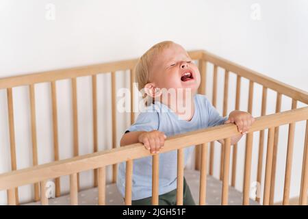Portrait of upset sad frustrated one year old baby boy getting hysterical standing in bed asking to pick him up, seeking attention of parents crying out loud. Child temper tantrum Stock Photo