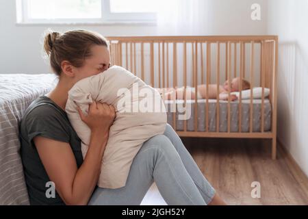 Depressed mother screaming with anger and desperation in pillow to overcome negative emotions, suffering postnatal depression because of lack of help and support with childcare routine Stock Photo