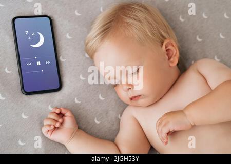 Closeup portrait of cute little boy watching sweet dreams in his bed falling asleep with music playing in app for baby sleep on phone lying next to his head. Happy maternity and technology Stock Photo