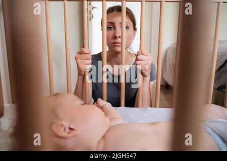 Young frustrated mother feels imprison while takes care of her infant and suffering from postnatal depression. Depressed woman looking at camera while her little toddler baby sleeping in his bed Stock Photo