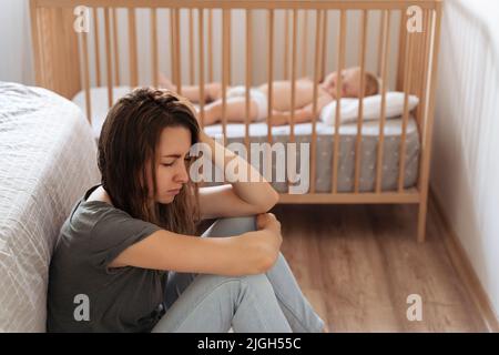 Young tired mother suffering from postnatal depression sitting on floor next to sleeping baby in crib with closed eyes. Feeling exhausted depressed demanding psychological help and support Stock Photo