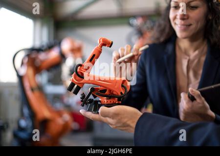 Close-up of man engineer holding model of industrial robotic arm and showing to collegue in factory. Stock Photo