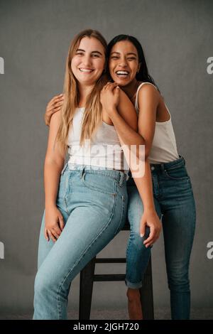 Two female friends posing in the studio sharing a stool Stock Photo