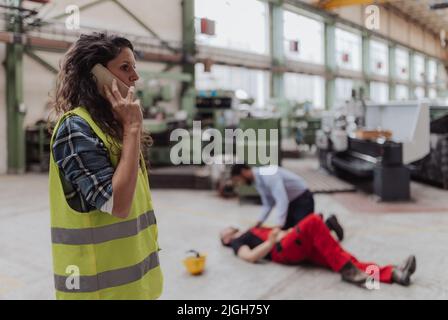 Woman is calling ambulance for her colleague after accident in factory. First aid support on workplace concept. Stock Photo