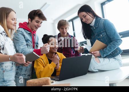 High school students sitting together at desk and using laptop and talking during break in classroom. Stock Photo