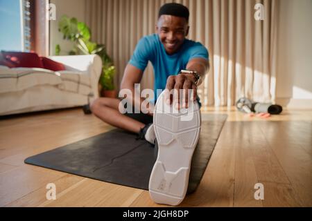 Young black African American happy male working out at home stretching his hamstring and legs legs, exercising in his living room on his yoga mat. Stock Photo
