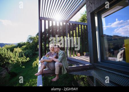 Young couple sitting and cuddling in hammock terrace in their new home in tiny house in woods, sustainable living concept. Stock Photo