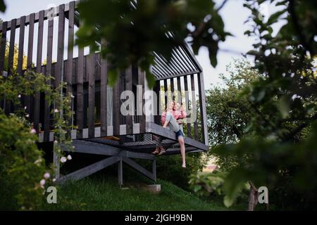 Young couple sitting and cuddling in hammock terrace in their new home in tiny house in woods, sustainable living concept. Stock Photo