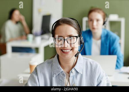 Portrait of young call centre agent in headphones and eyeglasses smiling at camera while working at office Stock Photo