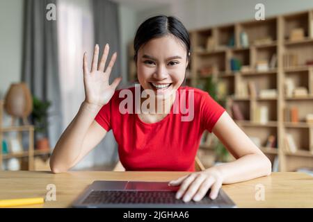 Smiling happy funny adolescent japanese girl typing on computer and waving hand at webcam in room or library Stock Photo