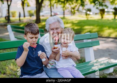 Great grandmother sitting on bench with her grandchildren and blowing soap bubbles together, generation family concept. Stock Photo