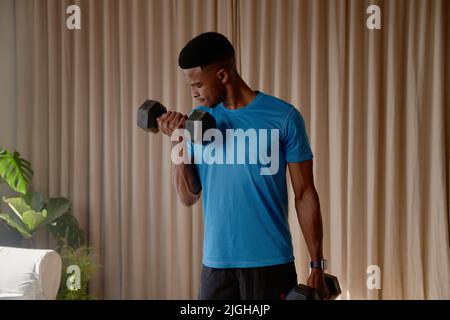 Front view of a Black African American young male exercising at home in his lounge lifting weights and dumbbells, bicep curls, keeping fit and healthy Stock Photo
