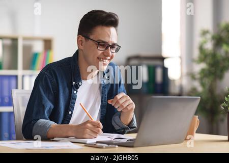 Handsome Young Businessman Using Laptop In Office And Taking Notes Stock Photo