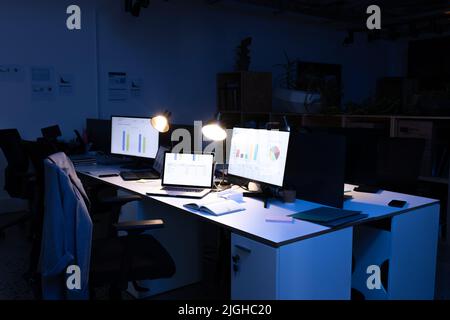 Computers and laptops with charts on screen and illuminated pendant lights on desk in office Stock Photo