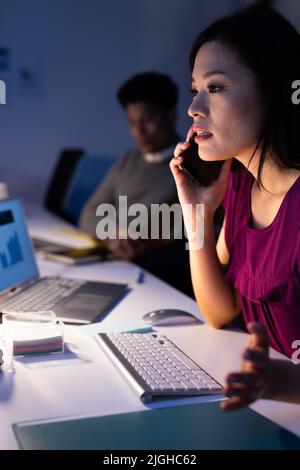 Asian businesswoman talking on smartphone and male colleague analyzing report at desk in office Stock Photo