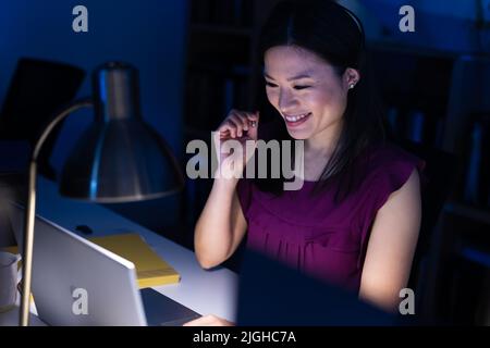 Smiling asian female customer service representative talking over headset and using laptop on desk Stock Photo